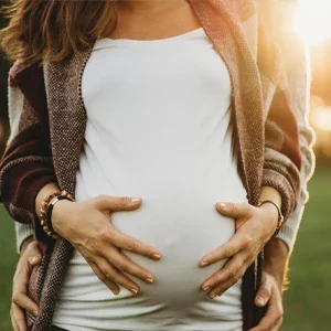 Hands gently resting on a pregnant abdomen outdoors, highlighted by warm natural light at sunset.
